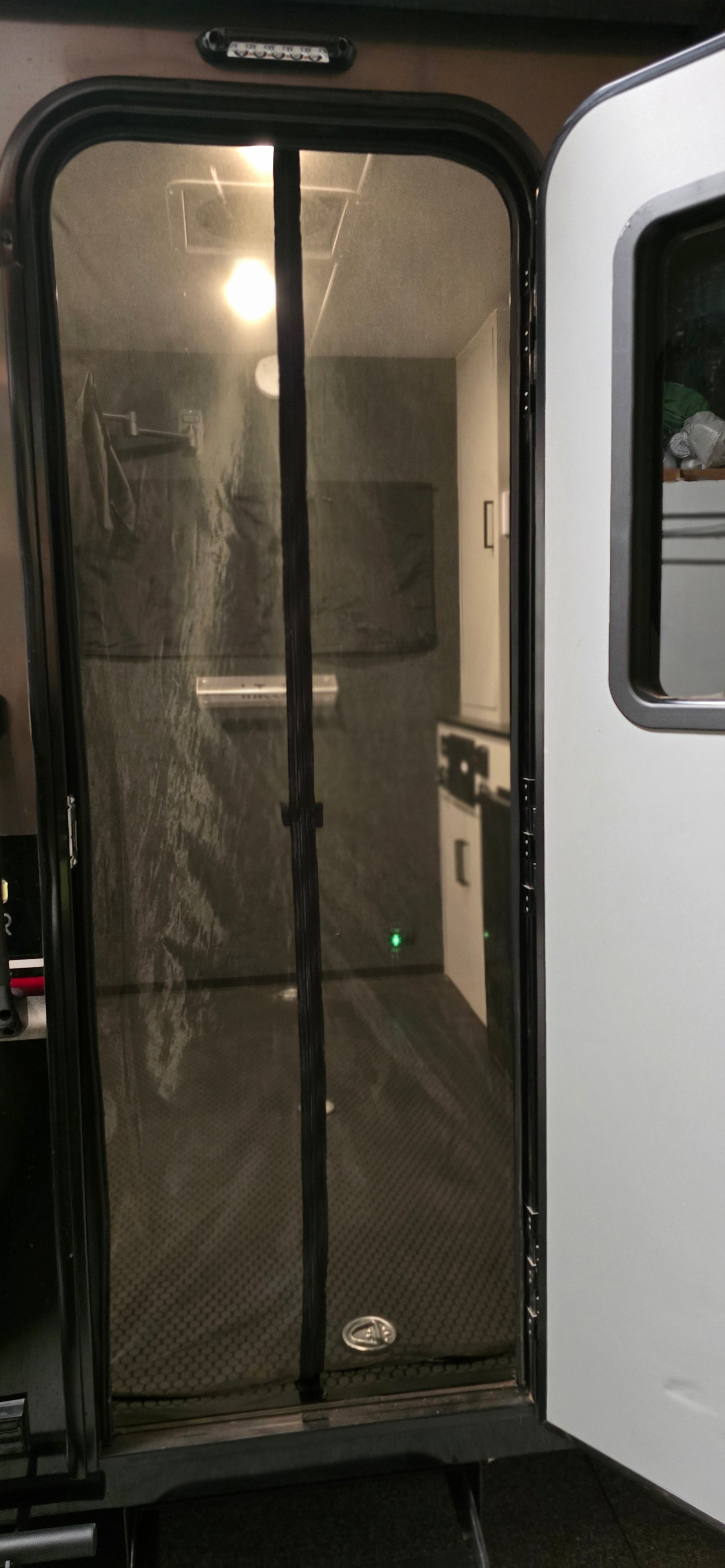 Peer through the Paha Que Custom Toy Hauler Mesh Door/Solid Room Divider into the camper, highlighting insect protection and revealing a cozy kitchenette with cabinets, lights, and a dark rubber floor mat.