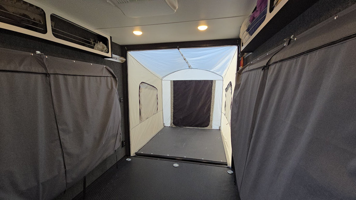The interior of a camper or RV is shown with gray curtains on both sides and an attached, enclosed tent area at the rear, visible through the open rear door. Overhead lights are illuminated and there are storage compartments above. The setup features the Toy Hauler Patio Enclosure Ramp Tent by PahaQue Wilderness, which is custom-built to fit all Toy Hauler trailers and provides versatile options for use as either a ramp tent or an outdoor patio space, enhancing comfort and functionality.