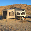 A Toy Hauler Patio Ramp Tent by Paha Que Custom, designed for Intech Discover Trailers, is set up on rocky desert ground near a white pickup truck, with dry hills and sparse brush under a clear blue sky in the background.