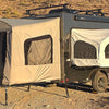 A black Intech Discover camper trailer equipped with two extended canvas pop-out tents and a PahaQue Custom Toy Hauler Patio Ramp Tent is parked on rocky desert terrain, with brown hills in the background.