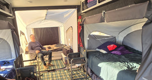 A man smiles inside a spacious camper with two elevated beds, checkered floor mat, camping chairs, and bedding as sunlight pours through an open PahaQue Wilderness Custom Toy Hauler Patio Ramp Tent for outdoor adventures.