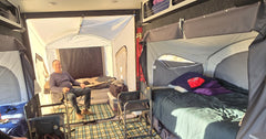 A man smiles inside a spacious camper with two elevated beds, checkered floor mat, camping chairs, and bedding as sunlight pours through an open PahaQue Wilderness Custom Toy Hauler Patio Ramp Tent for outdoor adventures.