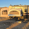 A Toy Hauler Patio Ramp Tent by Paha Que Custom is set up on an Intech Discover Trailer, resting on dry, rocky desert ground with shrubs and hills in golden sunlight. Camping supplies are placed near the tent entrance.