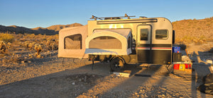 The Toy Hauler Patio Ramp Tent by Paha Que Custom for Intech Trailers is set up in a rocky desert at sunset, surrounded by sparse vegetation and hills, with outdoor gear beside it and weatherproof tent sections popped out.