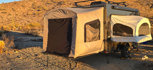A Paha Que Custom Toy Hauler Patio Ramp Tent for Intech Trailers is set up in a desert at sunset, with two pop-out tent sections surrounded by dry vegetation and hills.
