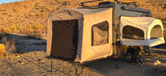 A Paha Que Custom Toy Hauler Patio Ramp Tent for Intech Trailers is set up in a desert at sunset, with two pop-out tent sections surrounded by dry vegetation and hills.