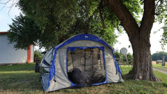 A Paha Que Custom Forest River R-Pod Trailer Side Tent for Enhanced Camping Comfort is set up under a tree by a gravel road, with a white and blue camper partly inside. Grass, gear, a metal shed, and wooden fence appear in the background.
