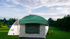 The Paha Que Custom 10x10 Side Mount Screen Room Tent for camping is set up on grass in front of a white camper trailer, with trees in the background and a partly cloudy sky above.