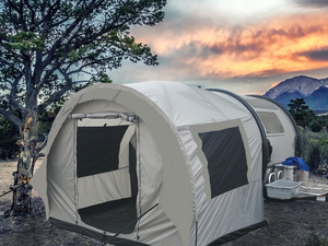 A Paha Que Custom NuCamp TAB 320 Trailer Side Tent for TAB and Boondock Trailers is set up outside near a tall tree, with camping supplies placed beside it and mountains plus a sunset sky in the background.