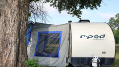 The Forest River R-Pod Trailer Side Tent by PahaQue Custom offers easy setup and enhanced camping comfort, pictured here beside a large tree on grassy terrain—an ideal spot to enjoy nature under blue skies.