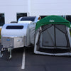 A Paha Que Custom 5x7 Trailer Side Tent Screen Room for Camping and Outdoor Adventures is set up next to a white teardrop trailer by a green-roofed canopy, with modern white buildings and landscaped greenery in the background.