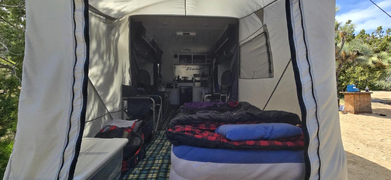 View from the entrance of a Paha Que Custom Toy Hauler Patio Ramp Tent for Intech Trailers, showing bedding, chairs, and camping gear inside; trees and a picnic table are visible outside.
