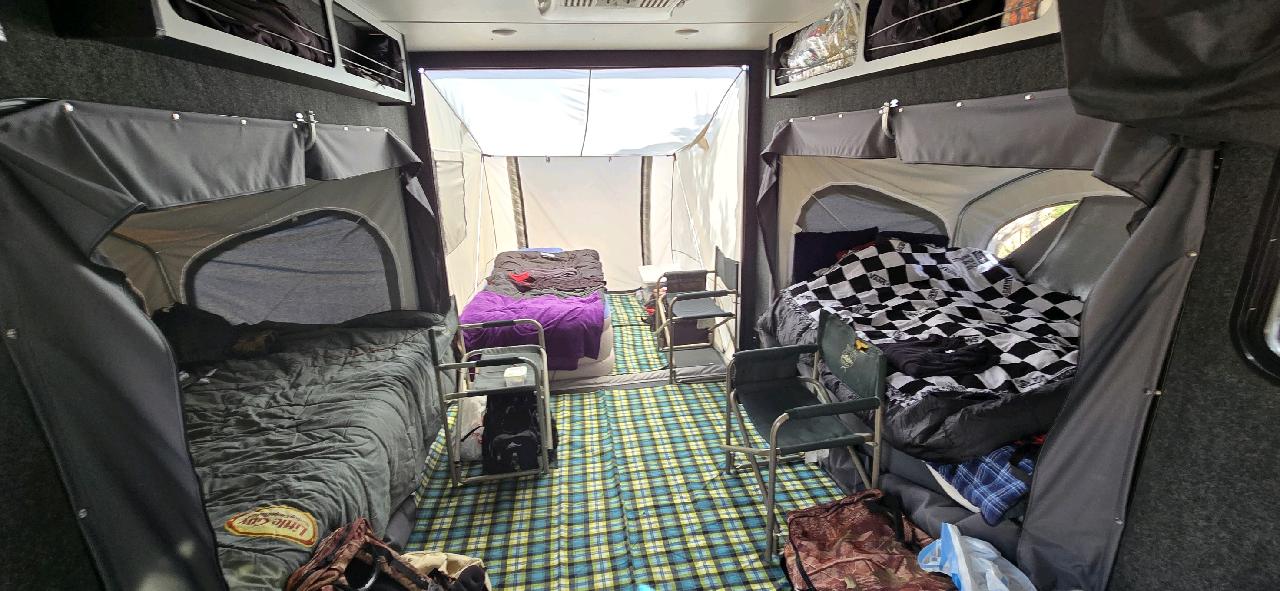 Experience comfort in any weather with the Toy Hauler Patio Ramp Tent by PahaQue for Intech Trailers—featuring three beds (one with a purple blanket, two side bunks), folding chairs, plaid carpet flooring, and a weatherproof design by Paha Que Custom.