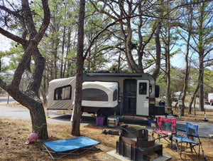 A pop-up camper is set up among pine trees at a campsite. Nearby, a PahaQue Wilderness Custom Toy Hauler Patio Ramp Tent for Outdoor Adventures, folding chairs, a fire pit, pink flamingo decor, and an outdoor mat complete the peaceful scene.