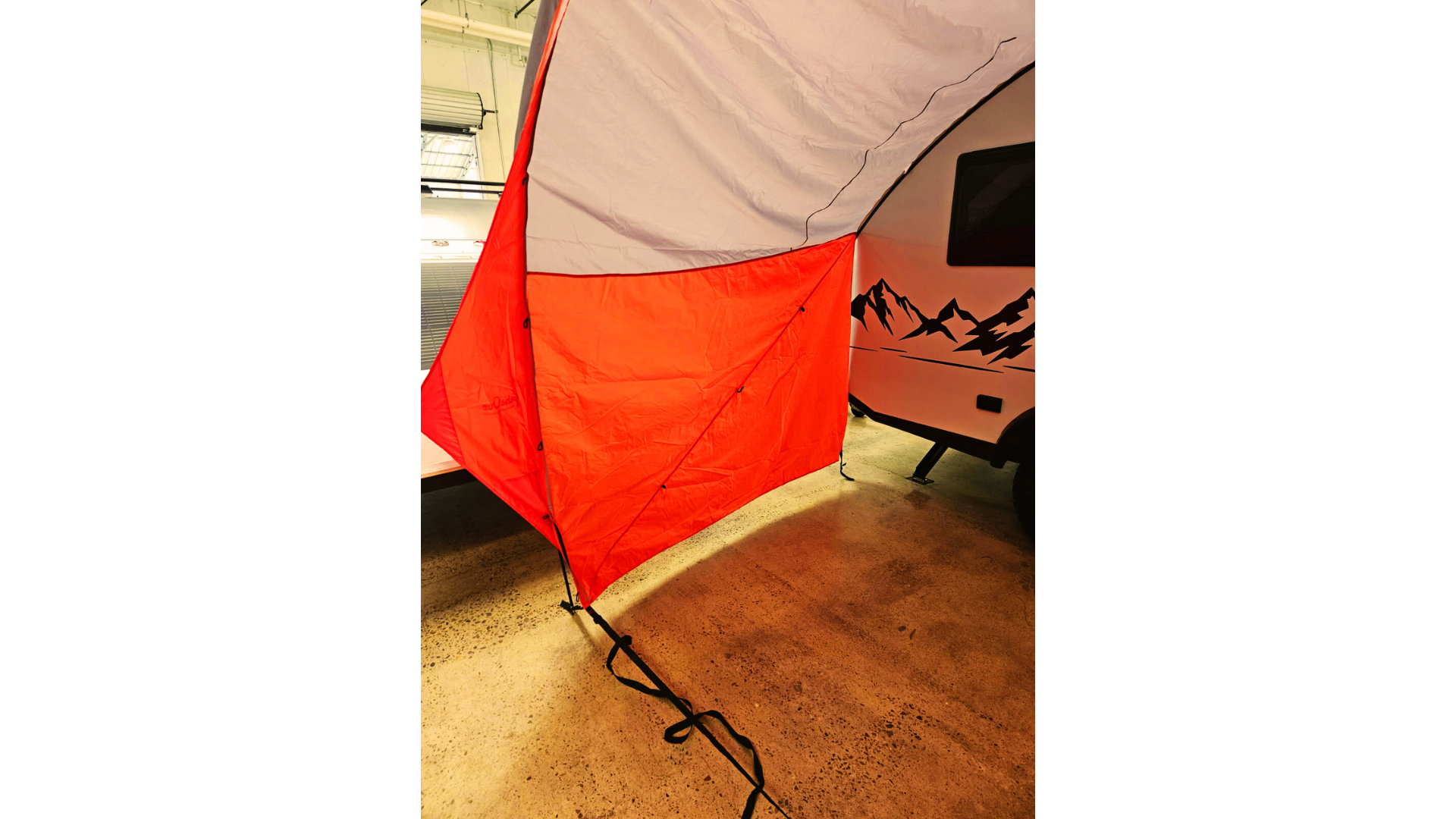 A NuCamp TAB 360 Trailer Awning by Paha Que Custom, in red and white, is attached to a camper trailer with mountain designs, providing waterproof indoor shelter over concrete. Straps keep the awning securely fastened to the ground.