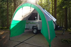A small camping trailer is parked in a forest, sheltered by a Paha Que Custom A Frame Trailer Awning for Aliner, Chalet, Rockwood, and Jayco trailers (12ft–18ft box sizes). Sunlight filters through the trees onto camping gear beside the trailer.