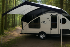The Alto Canopy by Paha Que Custom, a modern teardrop-style camper with a waterproof canopy, is parked on a forest path surrounded by tall trees and lush greenery.