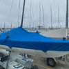 Several Soling sailboats are docked on land, their sails and cockpits protected by Paha Que Custom Soling Sailboat Storage Covers. Other boats and masts are visible in the marina under a cloudy sky.