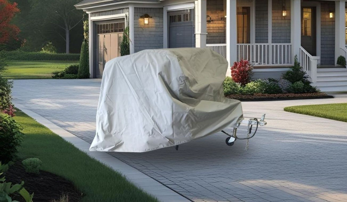 A vehicle is covered with the Ushi Chuck MAX Trailer Cover by Paha Que Custom, made for heavy-duty weatherproof protection, on a paved driveway in front of a gray house with white trim, porch, and landscaped yard with bushes and flowers.