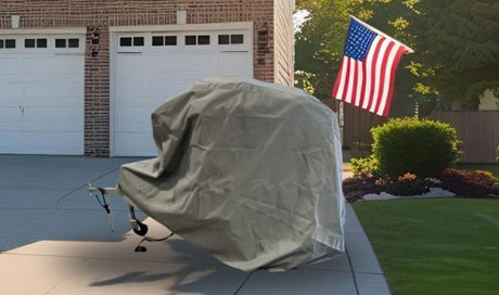 A trailer covered with the Ushi Chuck MAX Trailer Cover by Paha Que Custom, made from durable MagnaTuff fabric, is parked on a driveway in front of a brick house with white garage doors and an American flag waving nearby.