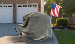 A vehicle with a gray Paha Que Custom Ushi Chuck MAX Trailer Cover by PahaQue Heavy Duty Weather Proof Protection is parked in a driveway near a garage. An American flag stands by the lawn, with trees and shrubs behind.