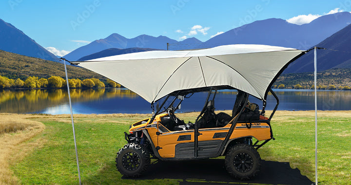 A PahaQue Wilderness All Purpose Portable Vehicle Awning for Cars, SUVs, Trucks, and Side by Sides is set up on grass near a mountain lake, surrounded by trees under a clear blue sky.
