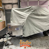 A small trailer with a durable Aero Teardrops Trailer Cover by PahaQue Wilderness made from MagnaTuff fabric is parked in a garage. A diamond-plate storage box is attached to the front, and the PahaQue Wilderness logo appears at the bottom.