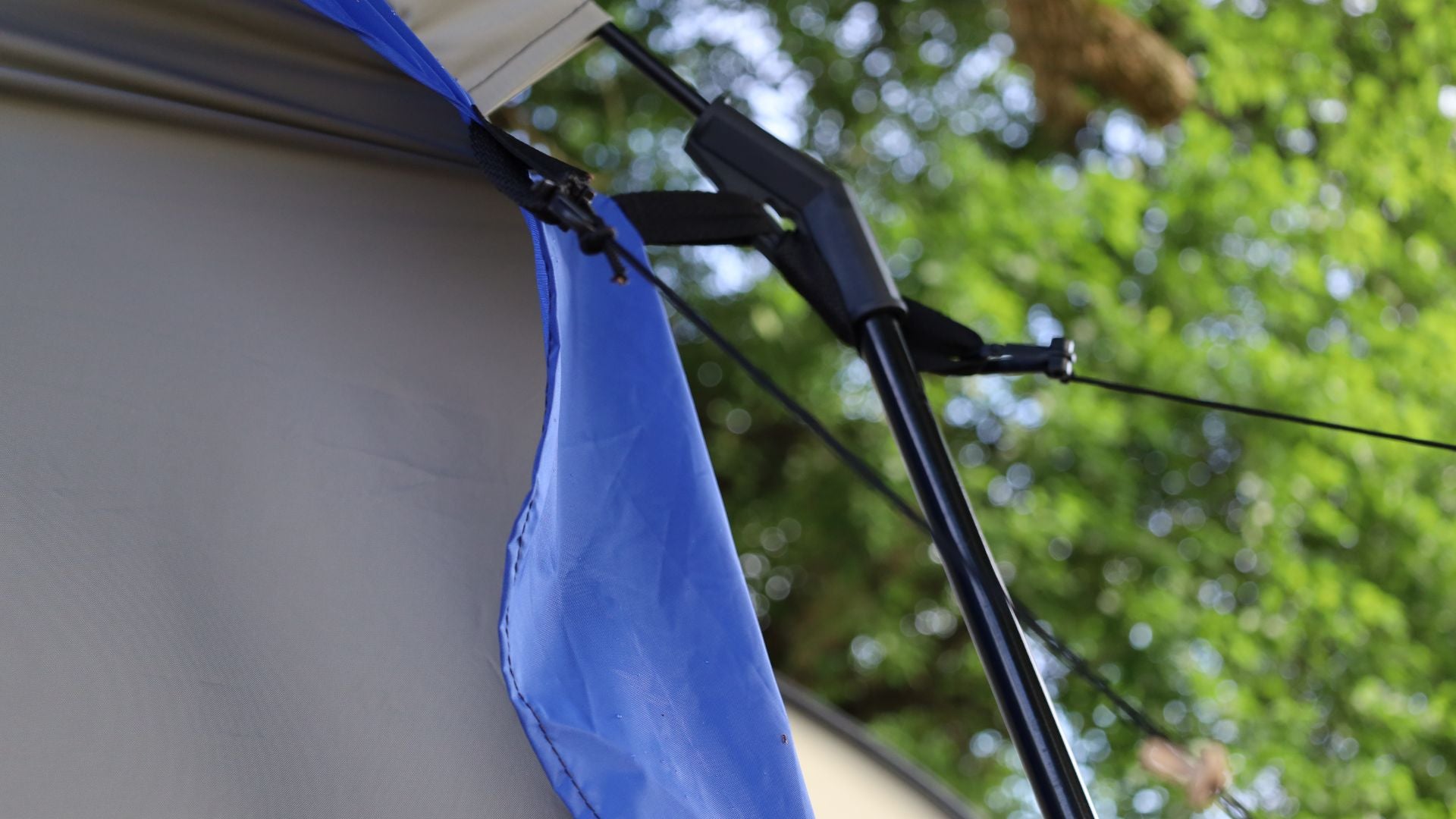 Close-up of the Forest River R-Pod Trailer Side Tent by PahaQue, showing a blue rainfly, tent pole, and guy line with black connector—camping gear from Paha Que Custom—against a backdrop of blurred green leaves and trees.