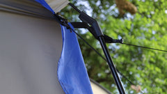 Close-up of the Forest River R-Pod Trailer Side Tent by PahaQue, showing a blue rainfly, tent pole, and guy line with black connector—camping gear from Paha Que Custom—against a backdrop of blurred green leaves and trees.