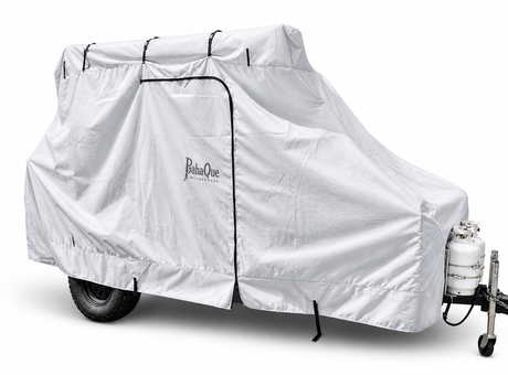 A white Boreas Campers Trailer Cover Premium Protection from Elements by Paha Que Custom is secured with straps and a side zipper over a small travel trailer with exposed tires and front propane tank, offering all-weather protection.