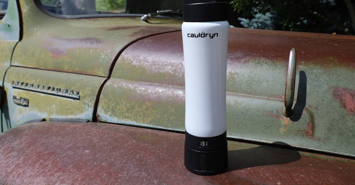 A PahaQue Wilderness Cauldryn Coffee Pro smart mug with a digital display rests on the rusty hood of an old, green truck parked outside.