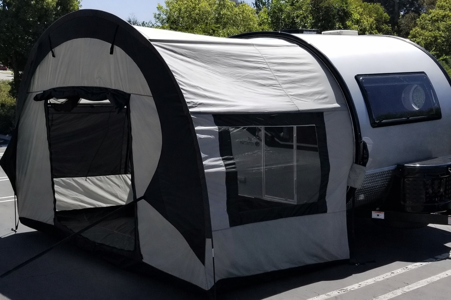 A gray and black NuCamp TAB 400 with a Paha Que Custom TAB 400 Trailer Side Tent by PahaQue is parked in a sunny lot surrounded by trees, with the attached awning and side tent set up.