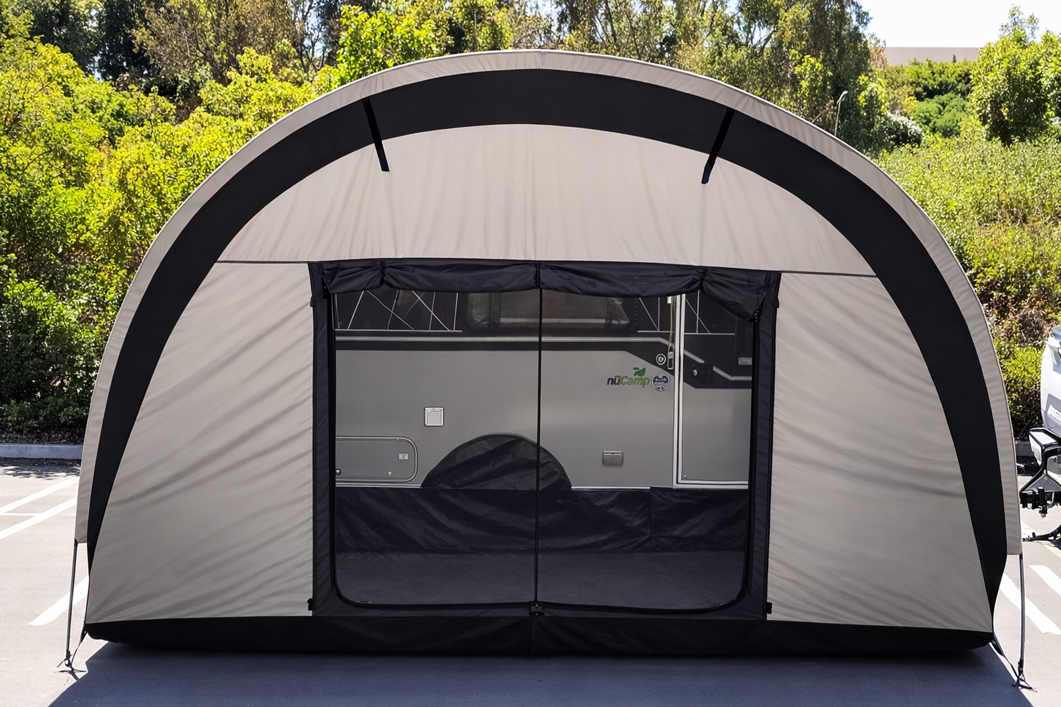 A beige and black portable canopy shelter shades a silver NuCamp TAB 400 Trailer Side Tent by Paha Que Custom for TAB Trailers in a parking lot, with trees and greenery visible in the sunlit background.