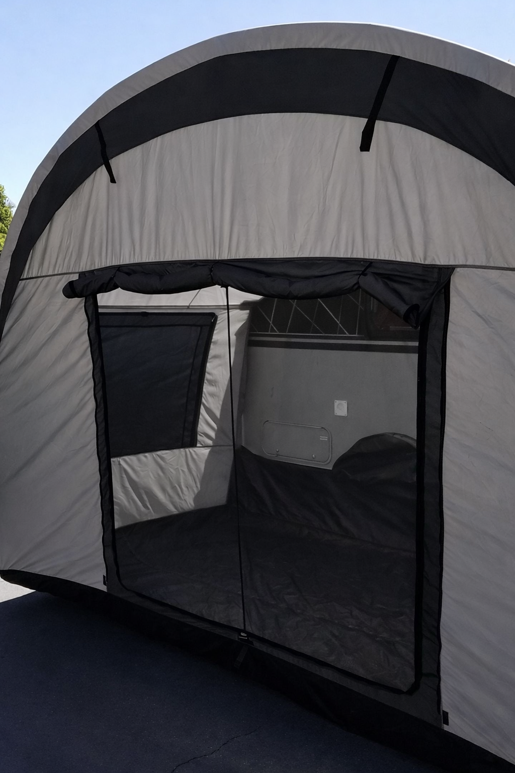 The Paha Que Custom NuCamp TAB 400 Trailer Side Tent for TAB Trailers is gray and black with a large zippered mesh door and window, shown partially open outdoors on a paved surface, ideal for use with TAB trailers.