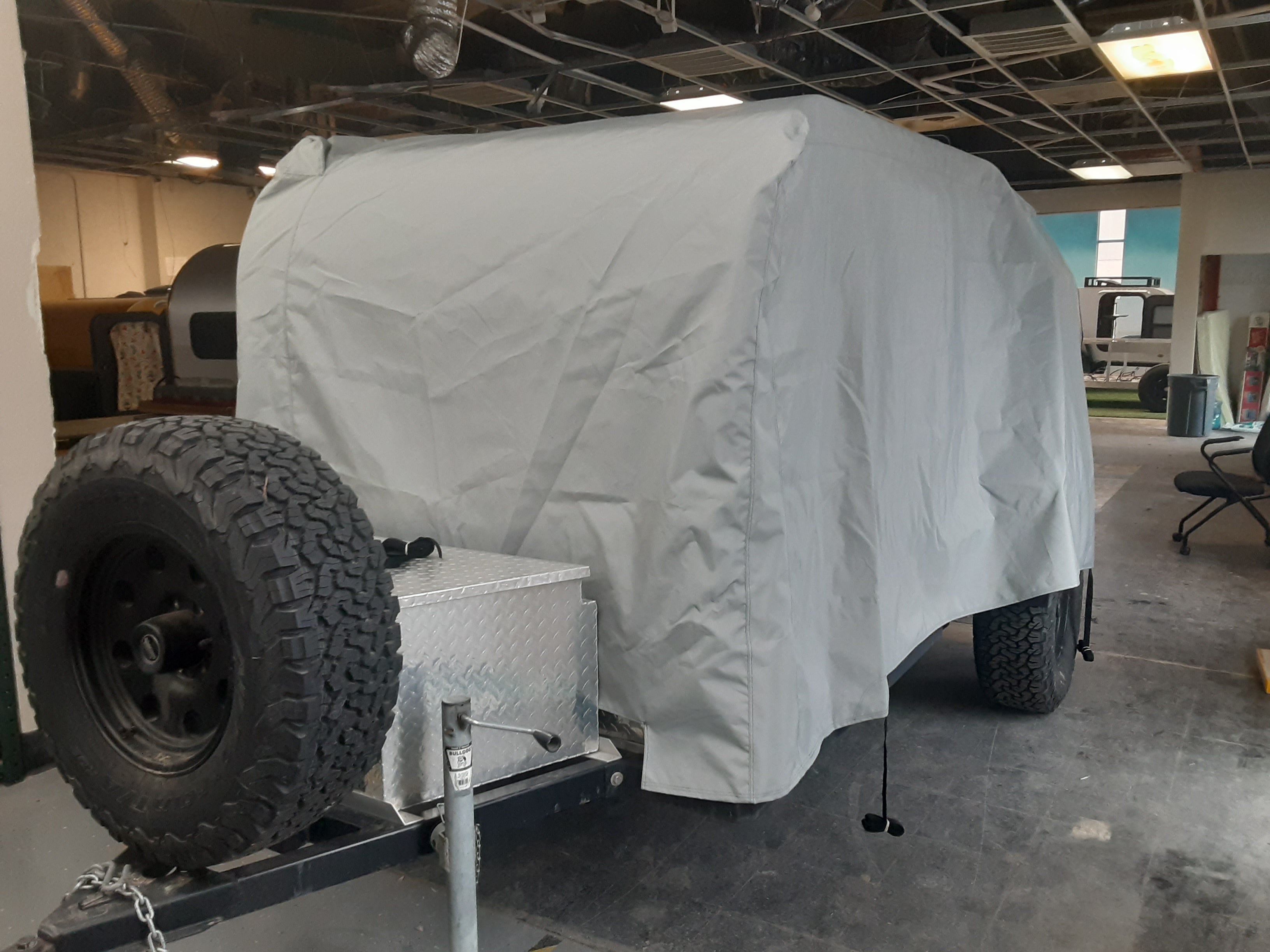 A PahaQue Wilderness Colorado Teardrops Canyonland Pinnacle Trailer Cover in gray MagnaTuff fabric shelters a small trailer with a large spare tire and metal storage box at the front, parked inside a building with a partially exposed ceiling.