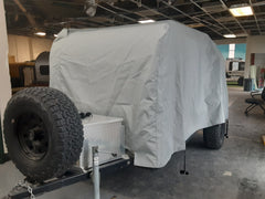 A PahaQue Wilderness Colorado Teardrops Canyonland Pinnacle Trailer Cover in gray MagnaTuff fabric shelters a small trailer with a large spare tire and metal storage box at the front, parked inside a building with a partially exposed ceiling.