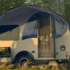 A small white and blue camper trailer features a Paha Que Custom Helio O Series Trailer Visor by PahaQue Outdoor Shelter Enhancement with string lights, its awning offering shelter among rocks, grass, and green trees in the background.