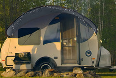 A small white and blue camper trailer features a Paha Que Custom Helio O Series Trailer Visor by PahaQue Outdoor Shelter Enhancement with string lights, its awning offering shelter among rocks, grass, and green trees in the background.
