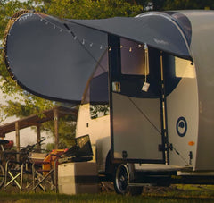 The Helio O Series Trailer Visor by Paha Que Custom is set up on grass, creating a modern outdoor shelter. Camping chairs and gear sit nearby, with string lights hanging from the curved awning as sunlight filters through the trees.