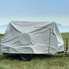A small trailer with a Hero Trailer Cover by Paha Que Custom, made from heavy-duty MagnaTuff fabric, is parked on green grass under a hazy blue sky, with mountains faintly visible in the background.