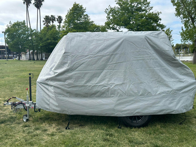A Squaredrop Trailer covered with the Paha Que Custom 6 x 13 Heavy Duty MagnaTuff Fabric trailer cover stands on grass in a park, with trees and palm trees visible under a partly cloudy sky.