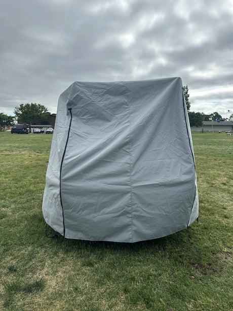 A Paha Que Custom Squaredrop Trailer Cover 6 x 13, made from heavy-duty MagnaTuff fabric, is set up on a grassy field under cloudy skies, with trees, people, and parked cars in the background.