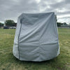 A Paha Que Custom Squaredrop Trailer Cover 6 x 13, made from heavy-duty MagnaTuff fabric, is set up on a grassy field under cloudy skies, with trees, people, and parked cars in the background.