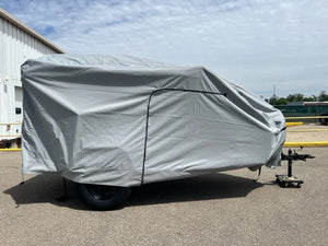 A Hiker Trailer is parked on pavement near industrial buildings, protected by a Paha Que Custom Hiker Trailer Covers Custom Fit Outdoor Protection. Only the trailer’s hitch and one wheel are visible under a partly cloudy sky.