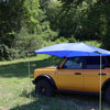 A yellow SUV is parked on grass with the PahaQue Wilderness All Purpose Portable Vehicle Awning attached, offering shade. Surrounded by green trees and bushes on a sunny day, its the ideal outdoor shade solution for cars, SUVs, and trucks.