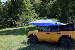 A yellow SUV is parked on grass with the PahaQue Wilderness All Purpose Portable Vehicle Awning attached, offering shade. Surrounded by green trees and bushes on a sunny day, its the ideal outdoor shade solution for cars, SUVs, and trucks.