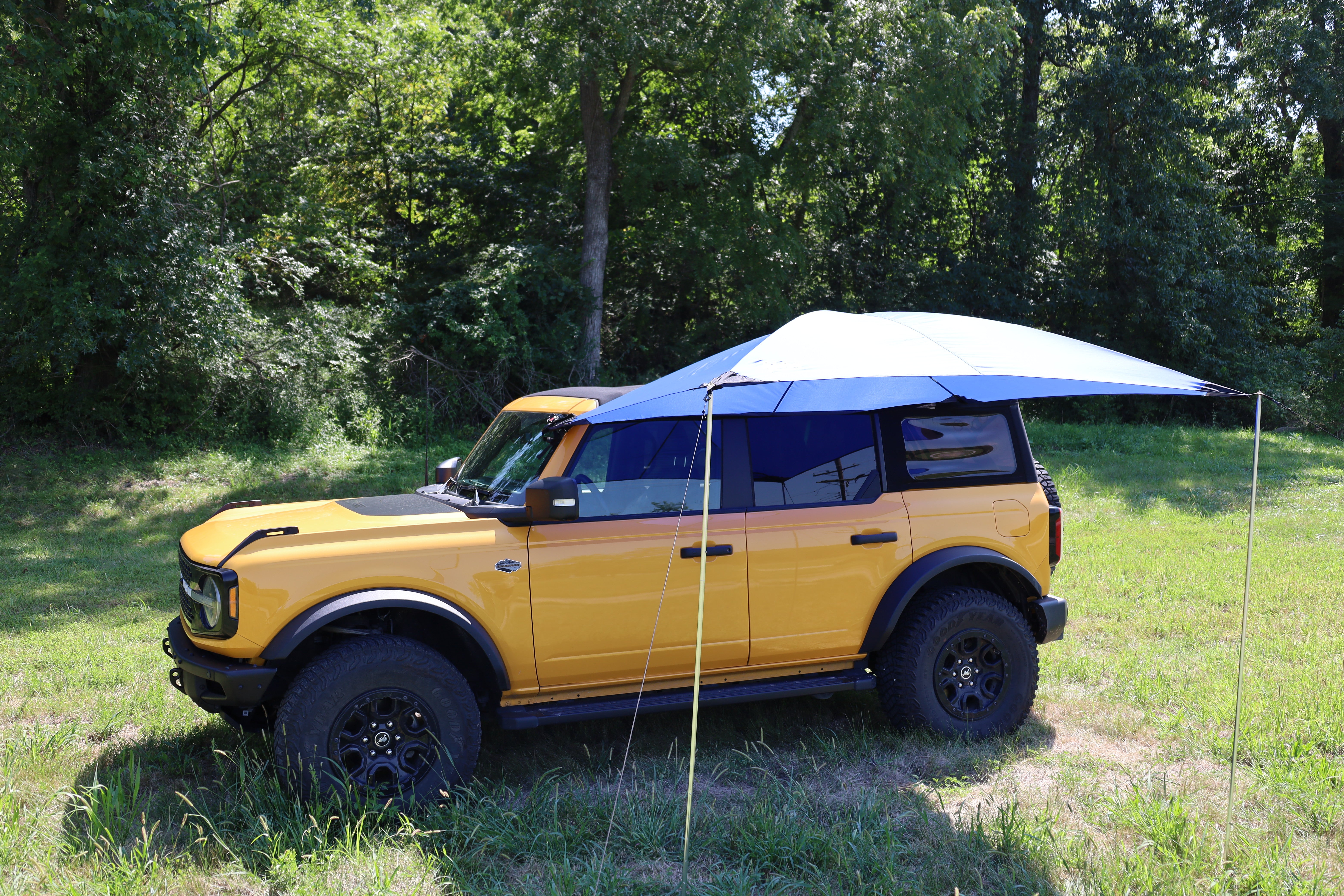 A yellow SUV is parked on grass by trees, equipped with the PahaQue Wilderness All Purpose Portable Vehicle Awning, creating ideal shade on one side—an excellent outdoor solution for cars, SUVs, trucks, and side by sides.