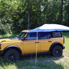 A yellow SUV is parked on grass by trees, equipped with the PahaQue Wilderness All Purpose Portable Vehicle Awning, creating ideal shade on one side—an excellent outdoor solution for cars, SUVs, trucks, and side by sides.