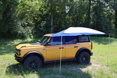 A yellow SUV is parked on grass by trees, equipped with the PahaQue Wilderness All Purpose Portable Vehicle Awning, creating ideal shade on one side—an excellent outdoor solution for cars, SUVs, trucks, and side by sides.