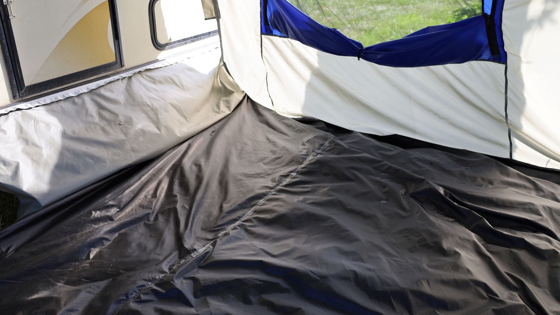 The photo shows the inside corner of the Forest River R-Pod Trailer Side Tent by Paha Que Custom, featuring a black floor, beige and white fabric walls, blue-topped window, and sunlight shining in—ideal for enhanced camping comfort.